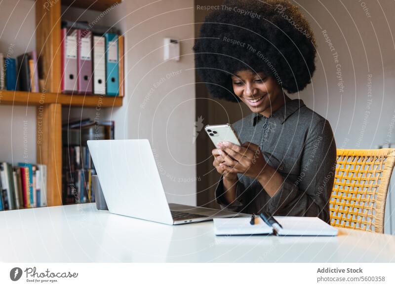 Fröhliche schwarze Frau, die in der Nähe ihres Laptops auf ihrem Smartphone surft Arbeit Textnachricht Browsen online freiberuflich Arbeitsplatz Internet