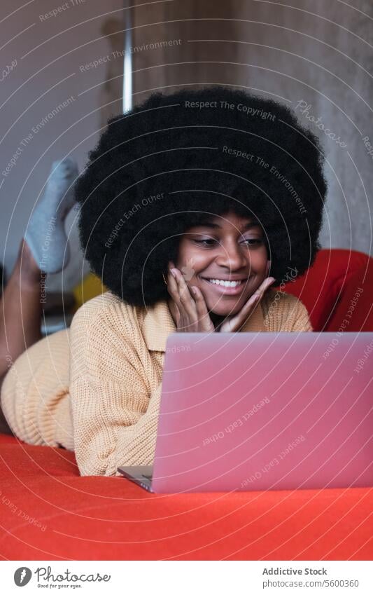 Lächelnde schwarze Frau tippt auf einem Laptop benutzend heimwärts Sofa Netbook Komfort sich[Akk] entspannen Browsen Tippen Afroamerikaner Afro-Look jung heiter