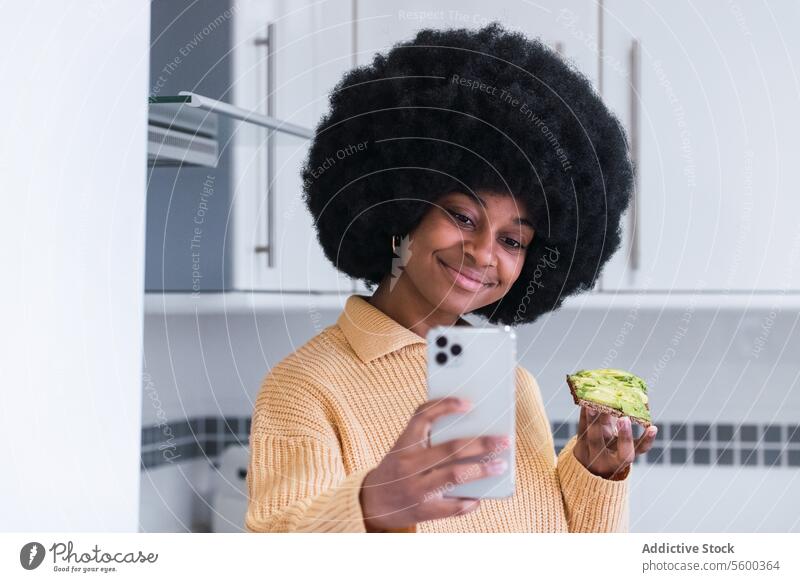 Lächelnde schwarze Frau macht Selfie mit Toast Frühstück Gesunder Lebensstil Küche Smartphone Selbstportrait Avocado benutzend Morgen Afroamerikaner Afro-Look