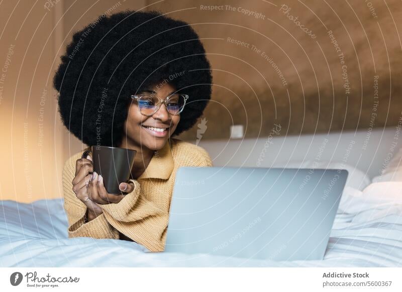 Lächelnde schwarze Frau mit Laptop und Kaffee im Bett benutzend ruhen sich[Akk] entspannen Wochenende Freizeit heimwärts Afroamerikaner Afro-Look zuschauen