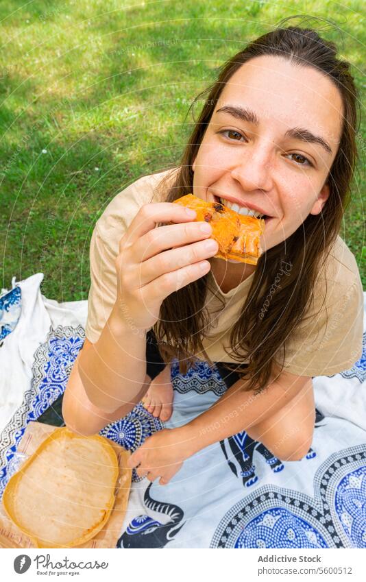 Junge Frau, die ein Picknick-Sandwich im Park genießt Belegtes Brot Essen Fröhlichkeit Erholung Decke sonnig im Freien heiter Genuss Snack Freizeit Sommer Gras