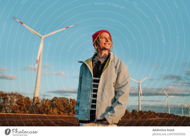 Lächelnde, alternative Frau in Jeansjacke, die von Windrädern wegschaut gelb Beanie Windkraftanlage nachhaltig Ökostrom Wegsehen heiter jung Erwachsener Porträt