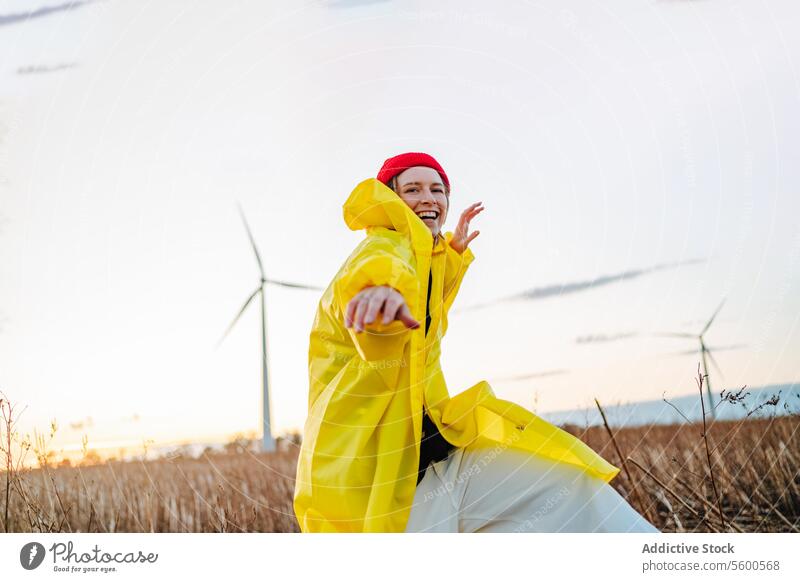 Lächelnde Frau in Gelb tanzt in der Nähe von Windkraftanlagen gelb Regenmantel rot Beanie Feld Sonnenuntergang Tanzen Freude pulsierend Natur