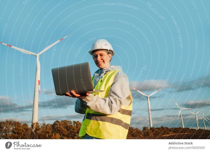 Ingenieurin mit Laptop im Windpark bei Sonnenuntergang Frau Windkraftanlage Fangvorrichtung Erneuerbare Energie Technik & Technologie Schutzhelm Lächeln