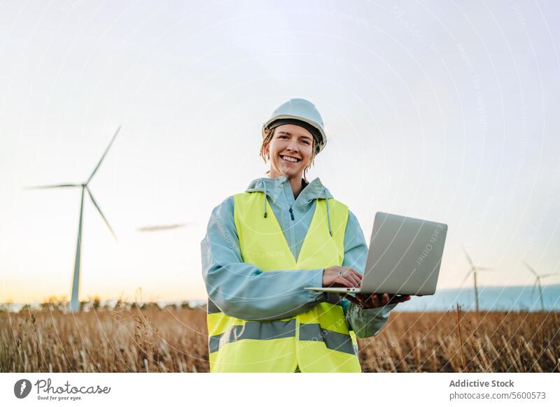 Ingenieur mit Laptop im Windpark bei Sonnenuntergang Frau Windkraftanlage nachhaltige Energie Technik & Technologie Feld Schutzhelm Warnschutzjacke Umwelt