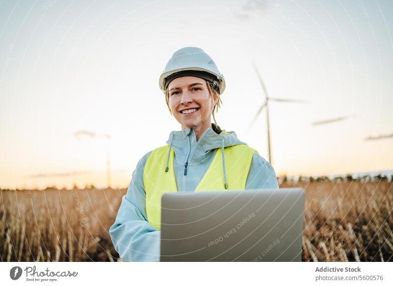 Lächelnder Ingenieur mit Laptop in der Nähe von Windkraftanlagen in der Abenddämmerung Frau Feld Sonnenuntergang Erneuerbare Energie Schutzhelm Sicherheitsweste