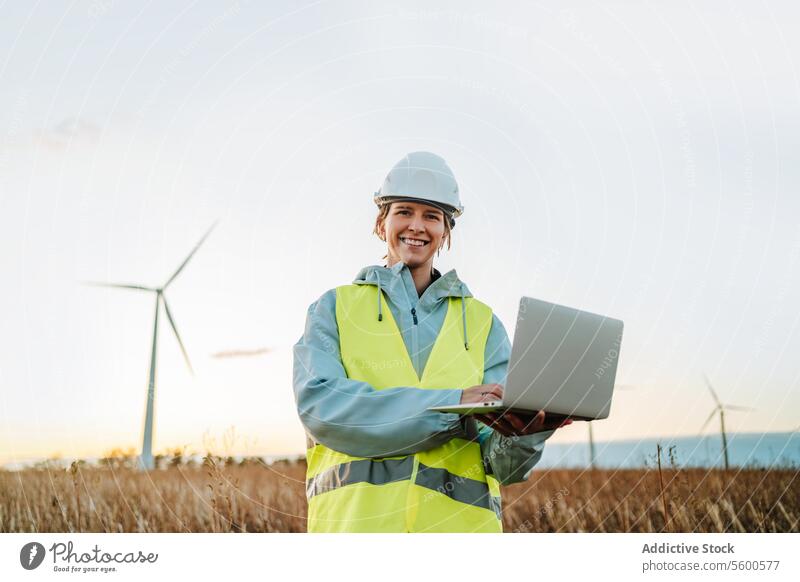Ingenieur, der mit seinem Laptop im Windpark bei Sonnenuntergang in die Kamera schaut Frau Windkraftanlage Feld in die Kamera schauen Fangvorrichtung