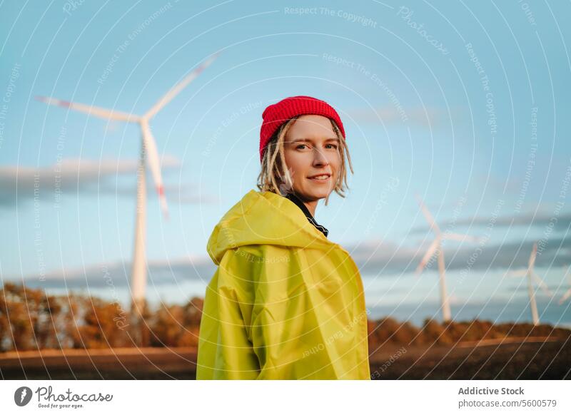 Lächelnde alternative Frau in gelber Jacke, die in die Kamera schaut, bei Windkraftanlagen Beanie nachhaltig Ökostrom heiter jung Erwachsener Porträt