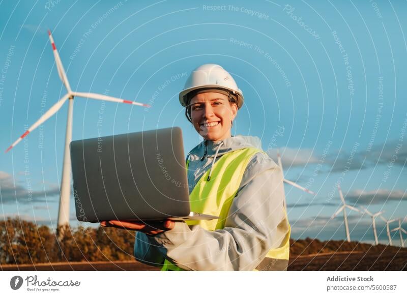 Ingenieurin, die bei Sonnenuntergang im Windpark mit Laptop in die Kamera schaut Frau Windkraftanlage Fangvorrichtung in die Kamera schauen Erneuerbare Energie