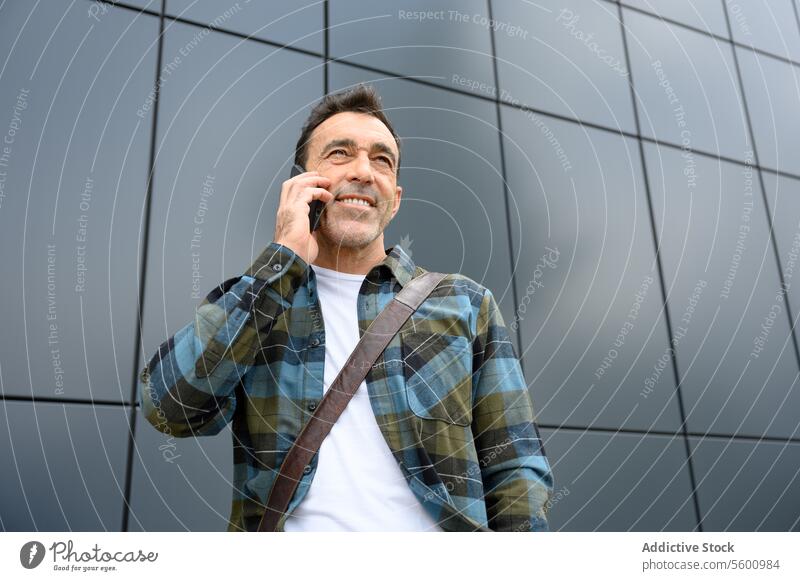 Fröhlicher junger Mann, der mit seinem Smartphone spricht männlich lässig Hipster Vollbart Straße reden Stil Wand modern Gespräch Telefonanruf Apparatur Lächeln
