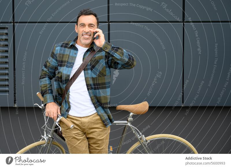 Fröhlicher Mann mit Fahrrad im Gespräch am Telefon männlich Smartphone Straße Hipster Stil lässig Großstadt gutaussehend urban Vollbart modern Lächeln Gebäude