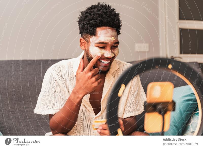 Lateinamerikanischer Mann trägt Gesichtsmaske während Videoblog-Sitzung auf Hautpflege heimwärts Schönheit Pflege Streaming lässig Spiegel Ringlicht Liege