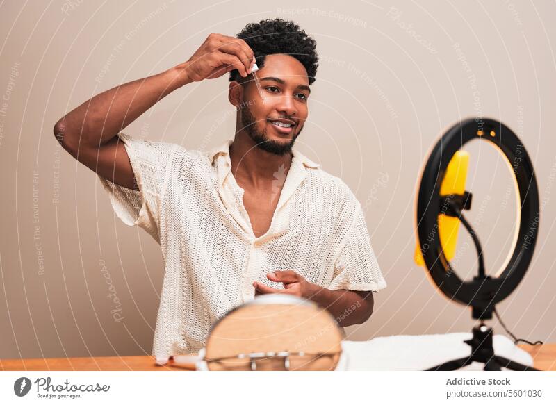 Lateinischer Mann, der ein Hautprodukt zu Hause mit Spiegel und Licht aufträgt Pflege Ringlicht Haarpflege Lächeln Frisur Kräusel Holztisch Hautpflege Routine