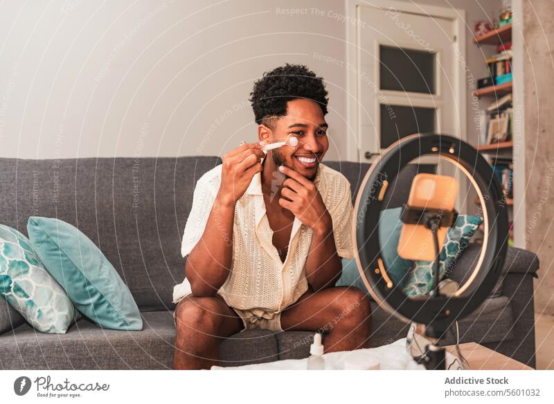 Ein fröhlicher junger Lateinamerikaner trägt in einem gut beleuchteten Wohnzimmer einen Gesichtsroller auf, der einen modernen Selbstpflege-Lebensstil für Video-Blog-Streaming widerspiegelt