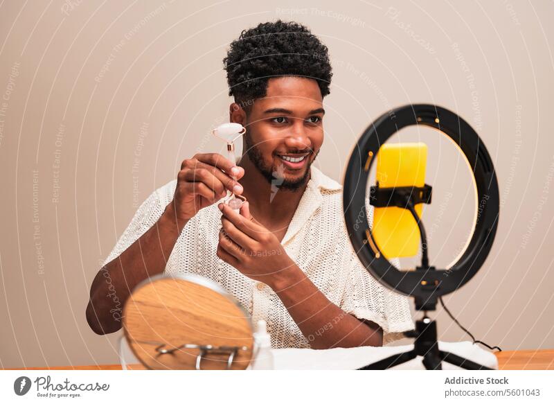 Lateinamerikanischer Mann lächelt bei der Aufnahme eines Videos mit Ringlicht mit einem Gesichtsroller Gesundheitswesen Haut Lächeln Smartphone Inhalt