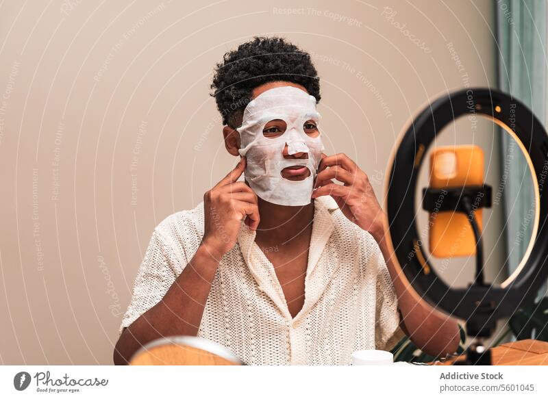 Junger lateinamerikanischer Mann, der eine Hautpflegemaske aufträgt und in den Spiegel schaut. Gesichtsmaske Schönheit Selbstfürsorge anwendend verwöhnend