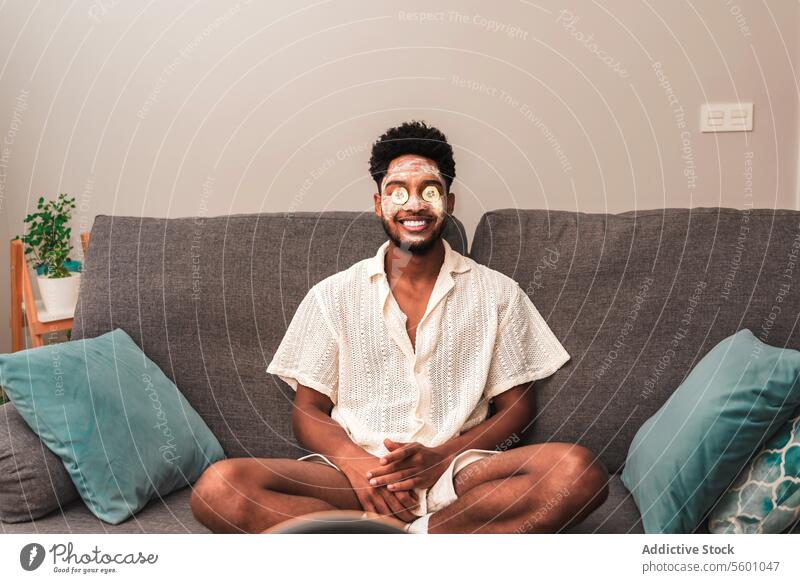 Lateinamerikanischer Mann entspannt sich zu Hause mit einer Gesichtsmaske Selbstfürsorge Hautpflege Erholung heimwärts Liege heiter Sitzen Wellness Schönheit