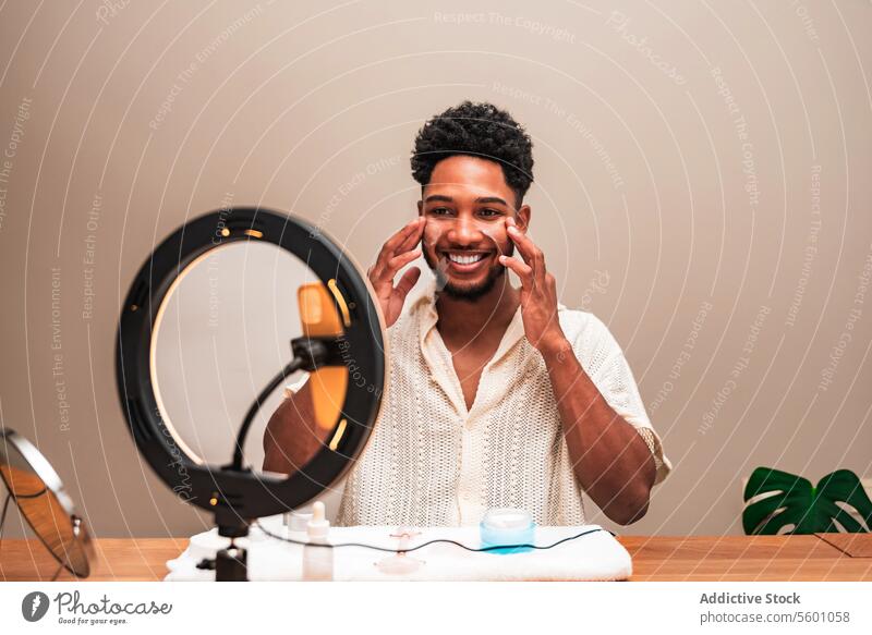 Junger lateinamerikanischer Mann beim Auftragen von Hautpflegecreme vor einem Ringlicht Sahne Spiegel Tisch Gesichtsbehandlung Produkt Pflege Routine Gesundheit