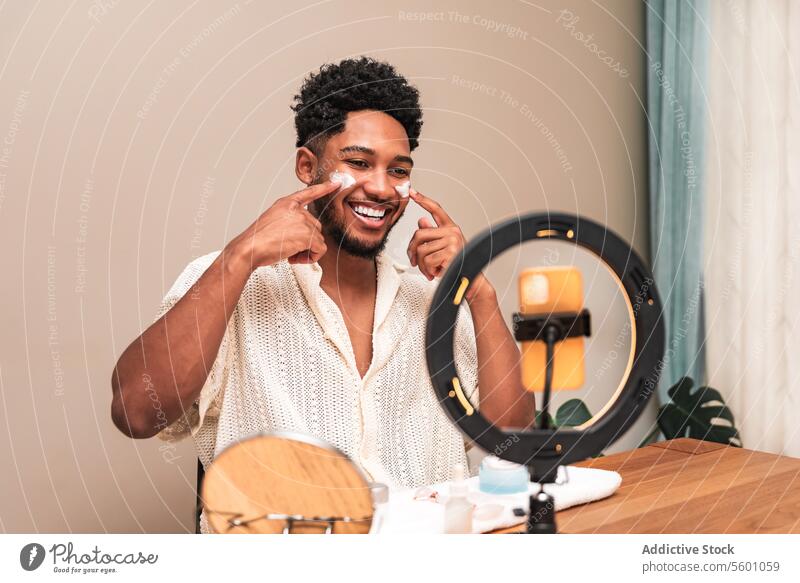 lateinamerikanischer Mann lächelt beim Auftragen der Hautpflege im Spiegel Routine Lächeln heimwärts Selbstfürsorge Schönheit Ringlicht Zeigen Wange Freude