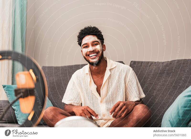 Lächelnder junger lateinamerikanischer Mann in Freizeitkleidung mit Gesichtscreme Sahne Hautpflege lässig Hemd Sitzen Liege heimwärts Ringlicht heiter