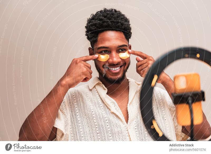 Junger lateinamerikanischer Mann beim Auftragen von Hautpflegeprodukten mit Gesichtspflaster vor Ringlicht jung Gesichtscreme Smartphone spielerisch