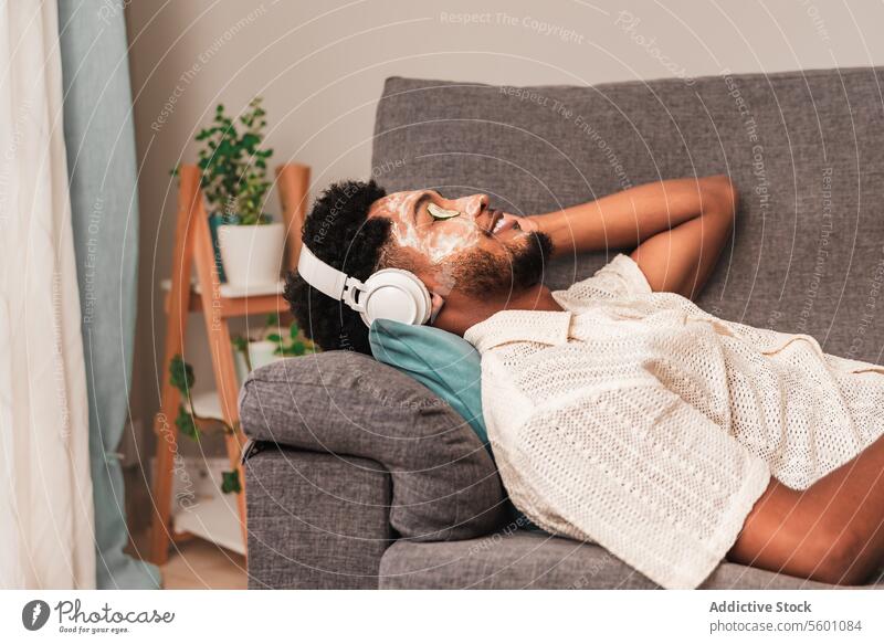 Junger lateinamerikanischer Mann genießt Musik auf Sofa mit Gesichtsmaske lehnen Sie sich zurück Liege Kopfhörer Augen geschlossen Erholung heimwärts