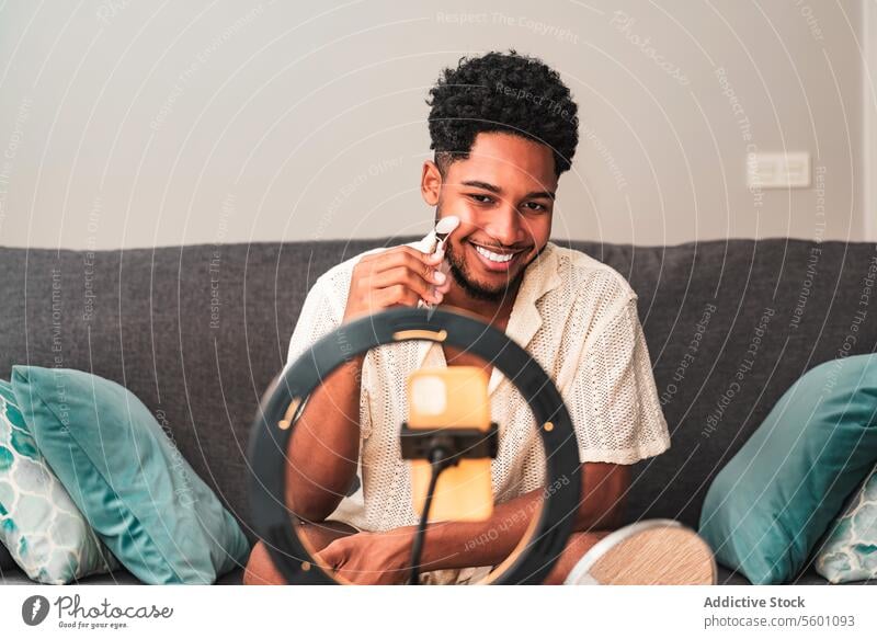 Ein fröhlicher junger Lateinamerikaner nimmt einen Videoblog zum Thema Hautpflege mit seinem Smartphone auf, das an einer Ringleuchte befestigt ist, und lächelt, während er zu Hause auf einer Couch einen Gesichtsroller benutzt.