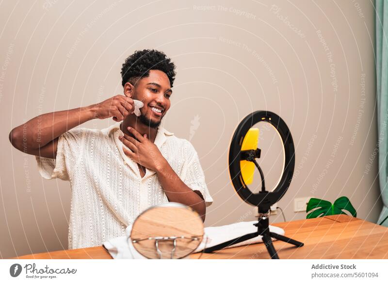 Junger lateinamerikanischer Mann, der ein Produkt zur Gesichtsmassage in einem Make-up-Setup für Streaming verwendet jung Tisch Ring Licht lässig Hemd