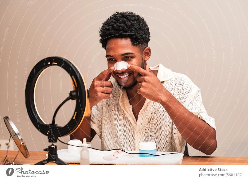 lateinamerikanischer Mann, der mit einem Lächeln seine Hautpflege aufträgt Nasenleiste Selbstfürsorge Ringlicht jung anwendend Pflege Routine Gesichtsbehandlung