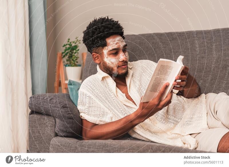 Lateinamerikanischer Mann, der sich zu Hause mit einer Gesichtsmaske entspannt und ein Buch liest Sofa lesen Hautpflege heimwärts räkeln sich[Akk] entspannen