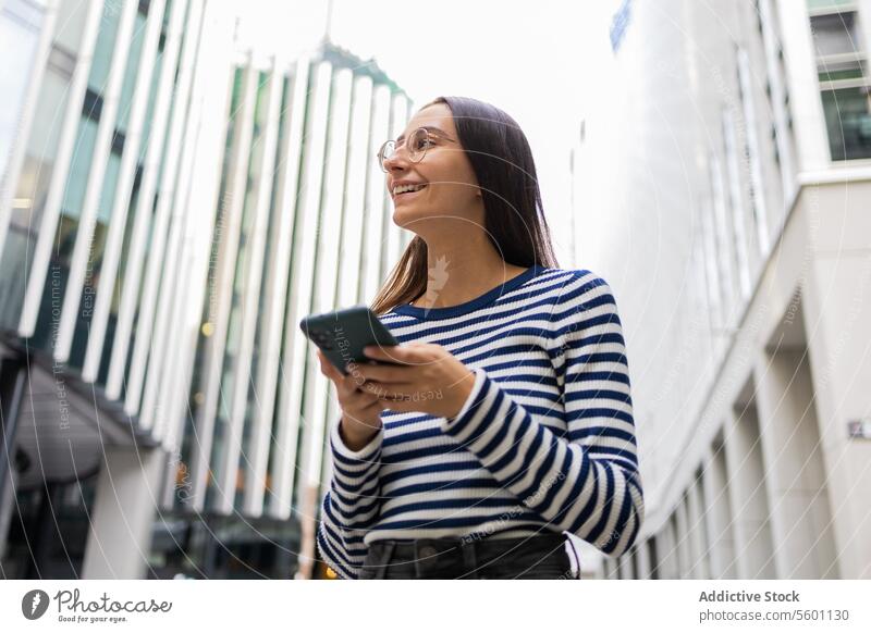 Lächelnde Frau mit Telefon auf einer Londoner Straße Smartphone moderne Gebäude gestreiftes Hemd jung Stadtleben Technik & Technologie urban lässig Mode