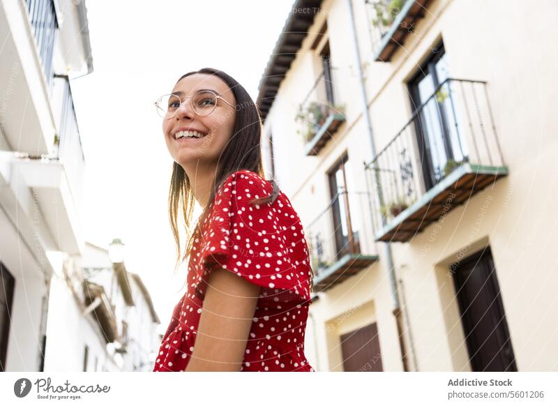 Niedriger Winkel der glücklichen Frau mit Brille, die gegen alte Gebäude in der Stadt wegschaut Tourist Lächeln Wegsehen heiter Freizeitkleidung Stehen Glück