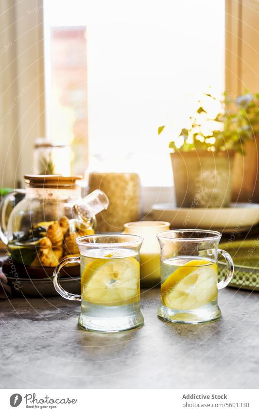 Gesundes Getränk mit Zitrone und Ingwer in gläsernen Teetassen am Küchentisch mit Tassen, Kerze und Kräutern am Fenster Hintergrund mit natürlichem Licht. Wärmende Heißgetränke mit Zitrusfrüchten. Vorderansicht.