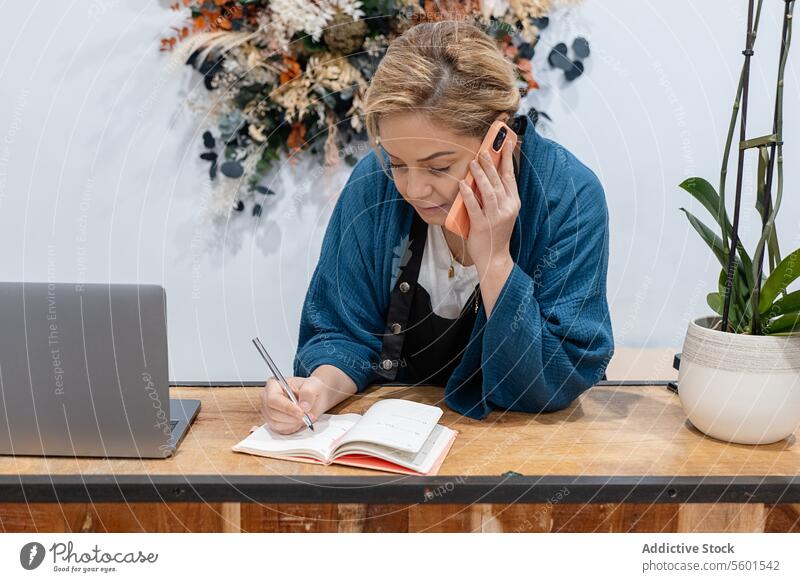 Geschäftsfrau, die ihren Terminkalender mit Notizen und Laptop organisiert und über ihr Smartphone spricht Frau schreibend Planer Schreibtisch hölzern Zeitplan