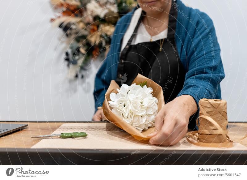 Anonyme Person, die einen Strauß mit weißen Blumen und Papier bastelt Blumenstrauß Handwerk umhüllen Tisch hölzern Garn Schere geblümt Ordnung Hobby Design