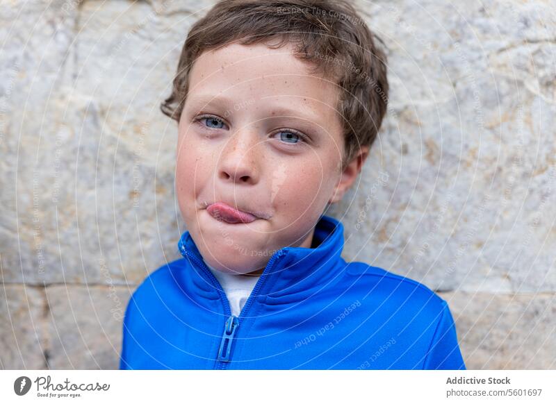 Blauäugiger Junge, der mit einem lustigen Gesicht in die Kamera schaut lustiges Gesicht Kind Humor spielerisch blau Jacke Stein Wand Ausdruck schelmisch