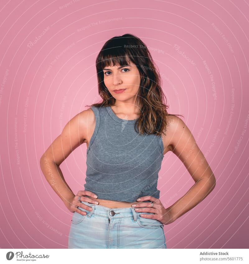 Studio-Porträt einer selbstbewussten Frau mit den Händen auf den Hüften, die in die Kamera schaut, vor einem rosa Hintergrund Errungenschaft Aktion aktiv