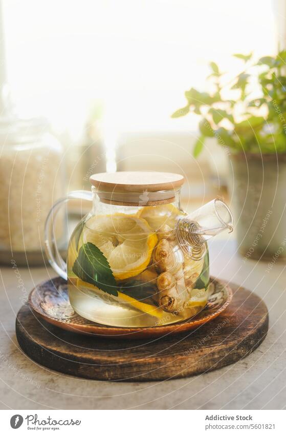 Glas-Teekanne mit Zitrone, Minze und Ingwer auf hölzernen Schneidebrett am Fenster Hintergrund mit natürlichem Licht. Gesunder Tee mit Vitamin C in der kalten Zeit. Vorderansicht.