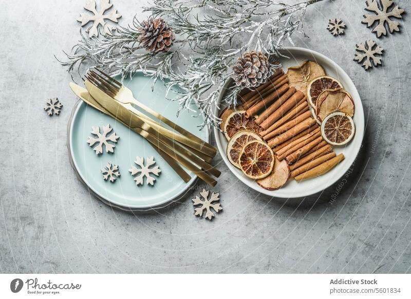 Weihnachtlicher gedeckter Tisch mit goldenem Besteck, hellblauem Teller, Zimtstangen, getrockneten Orangen und Äpfeln und Winterzweigen auf grauem Tisch.  Ansicht von oben.