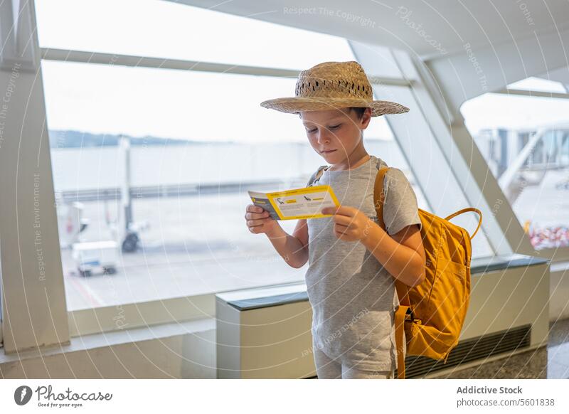 Porträt eines niedlichen nachdenklichen Jungen in Freizeitkleidung und Strohhut, der an der Fluggastbrücke steht und die Bordkarte im Flughafenterminal betrachtet
