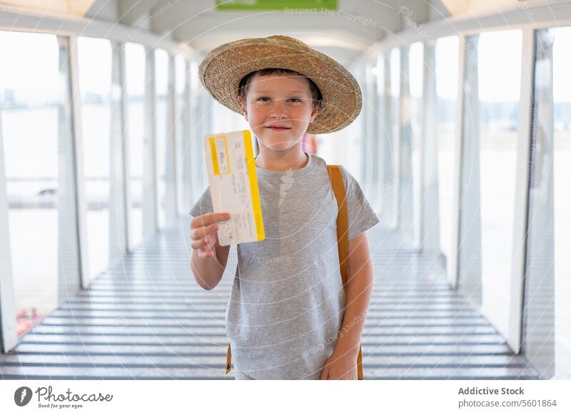 Porträt eines niedlichen lächelnden Jungen in Freizeitkleidung und Strohhut, der an der Fluggastbrücke steht und seine Bordkarte im Flughafenterminal zeigt