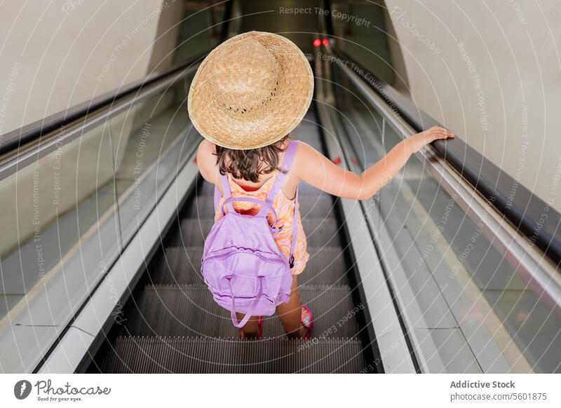 Hochwinkliges Ganzkörperbild eines anonymen Mädchens mit Strohhut und lilafarbenem Rucksack, das allein auf einer Rolltreppe im Flughafen steht Hut Fliederbusch