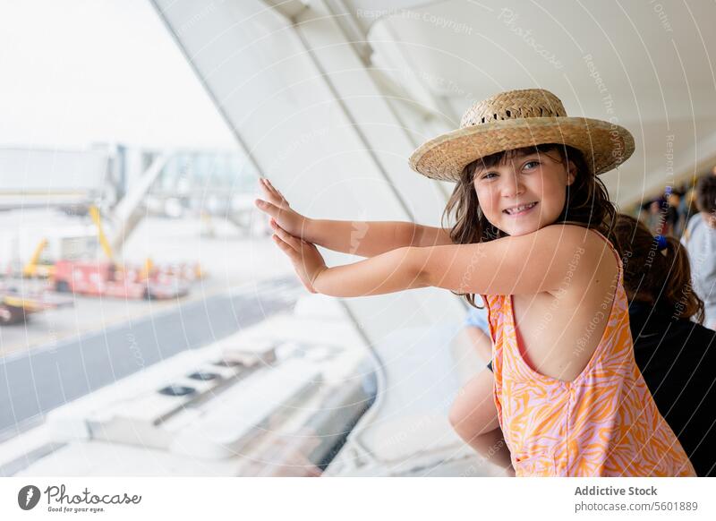 Seitenansicht einer gesichtslosen Reisenden mit Strohhut und Freizeitkleidung, die in die Kamera schaut, während sie ein Flugzeug auf der Rollbahn durch ein Fenster am Flughafen betrachtet
