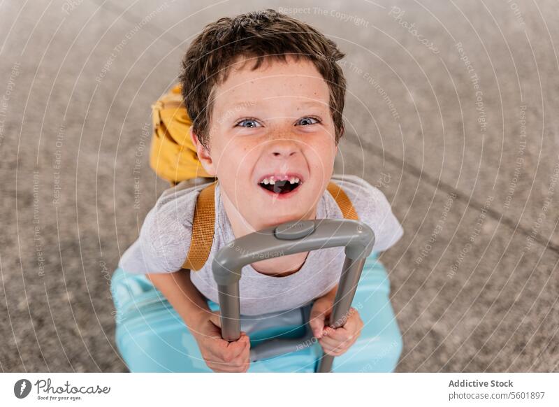Fröhliches Kind mit Gepäck von oben Junge spielerisch fehlender Zahn hoher Winkel heiter Urlaub Feiertag Porträt Lehnen Handwagen Flughafen Terminal Ausdruck