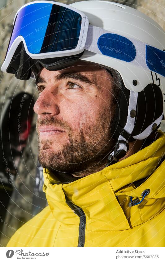 Selbstbewusster Skifahrer mit Helm und Skibrille schaut zur Seite Mann Vollbart gelb Jacke Schutzhelm Schutzbrille Nahaufnahme nachdenklich Blick Entfernung