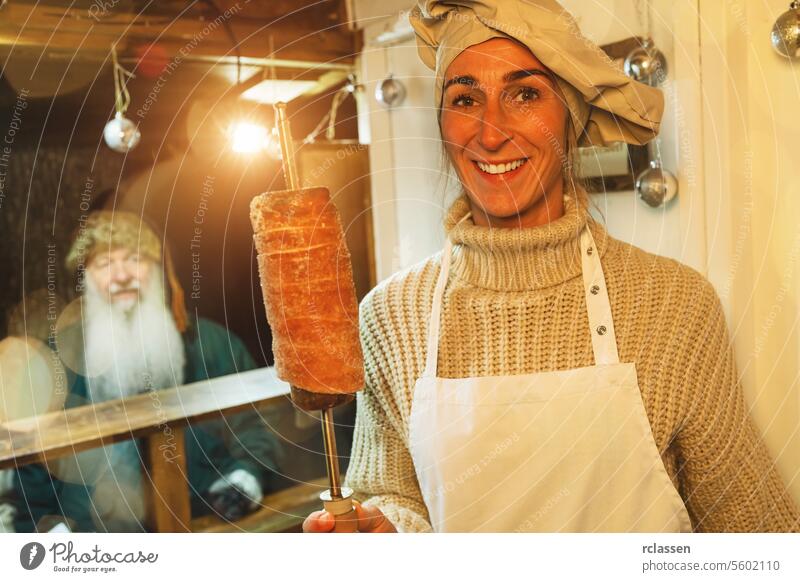 Lächelnde Verkäuferin mit Trdelnik, Kurtoscalacs oder Baumstriezel an einem Spiralstab in einem Stand auf dem Weihnachtsmarkt Vollbart alter Mann Dezember