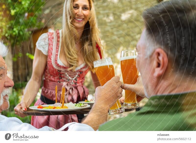 Kellnerin in Tracht, Dirndl, die in einem bayerischen Biergarten Essen serviert, im Hintergrund Menschen, die essen und trinken und mit Bierhumpen anstoßen alt