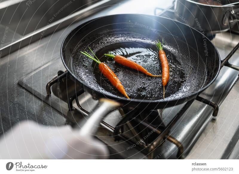 Hand schwenkt Karotten in einer geölten Pfanne auf einem Gasherd in einer professionellen Küche in einem Restaurant. Luxus-Hotel Kochen Konzept Bild. Gemüse