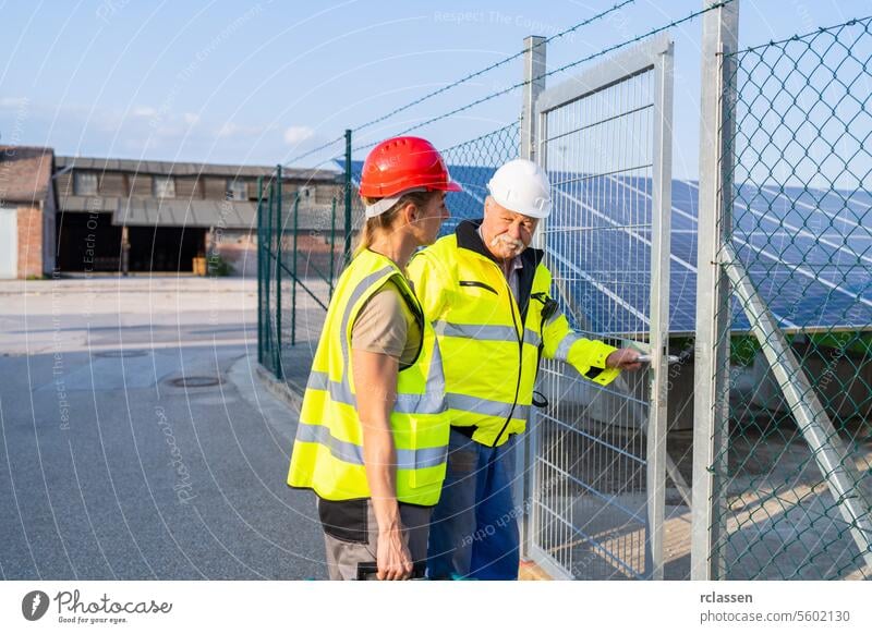 Zwei Ingenieure in Warnwesten inspizieren ein Solarmodulfeld, das durch ein Anlagentor betreten wird Besichtigung prüfen Solarmodulanlage Gate Sicherheitshelme