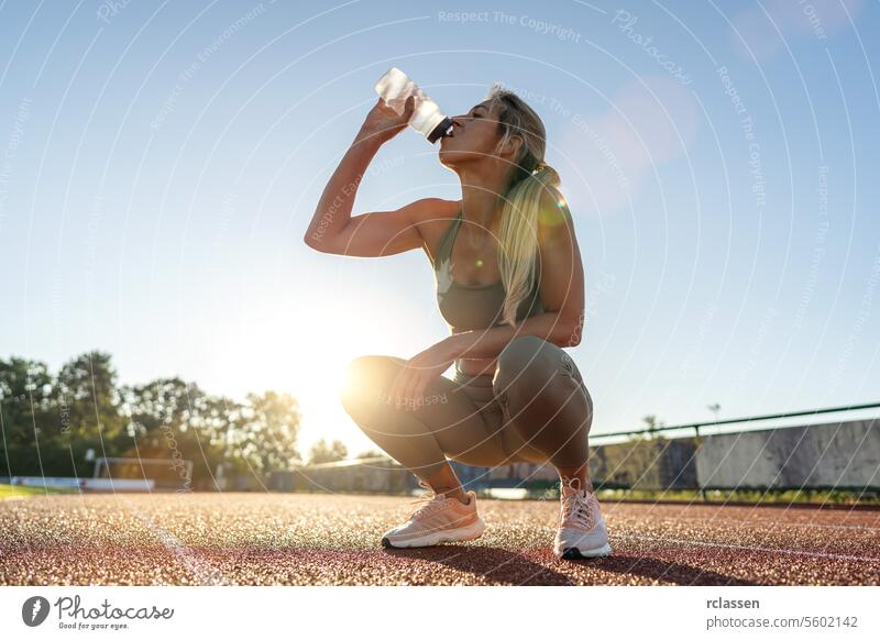 Frau trinkt Wasser, während sie bei Sonnenuntergang auf einem Sportplatz hockt Trinkwasser Hydratation Leichtathletik Fitness Athlet Sportbekleidung Gesundheit